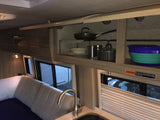 Inside an RV rental, showing the kitchen area with sink, full cabinets of cookware/dishes, and a bed. A warning label about glass/fire is visible.
