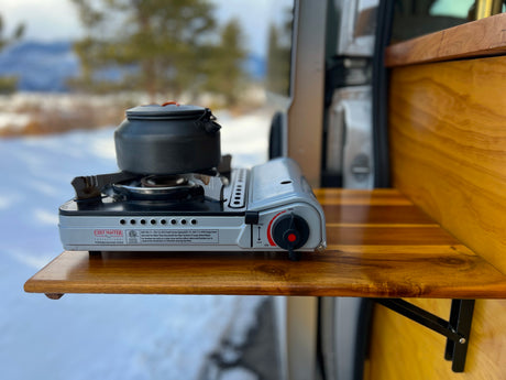Winter RV rental cooking: Portable butane stove with a kettle on a wooden counter, outdoors in a snowy mountain setting.
