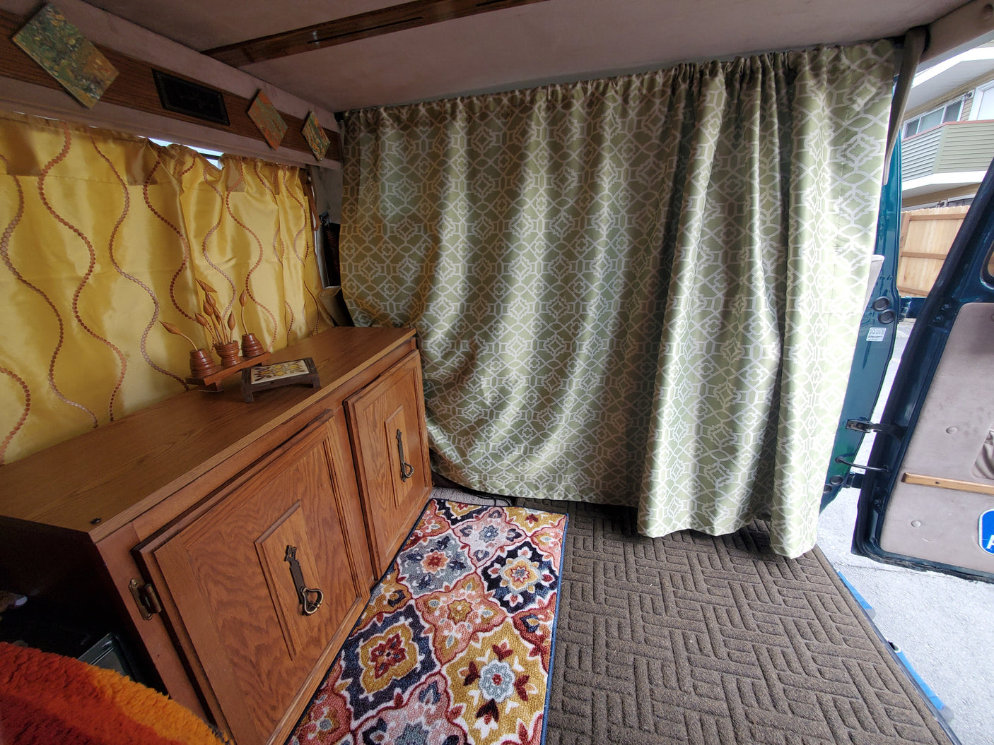 Inside a cozy RV rental: yellow wavy curtains, a wooden cabinet, and a green geometric curtain separating the space. Colorful rugs add warmth.