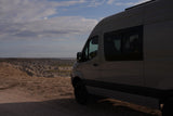 This grey RV rental camper van overlooks a vast, rugged badlands landscape beneath a cloudy sky, ready for adventure.