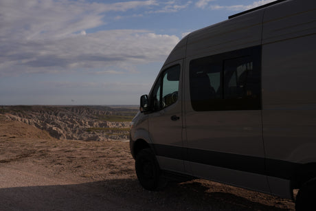 This grey RV rental camper van overlooks a vast, rugged badlands landscape beneath a cloudy sky, ready for adventure.