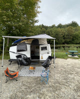 This rv rental travel trailer, with awning & chairs on a rug, is set by a lake for camping.