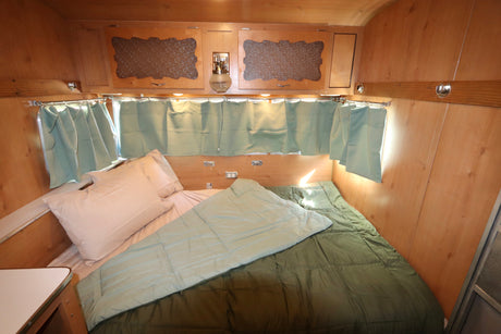 Comfortable RV rental sleeping area with natural wood paneling, a bed, green comforters, and vintage overhead cabinets.