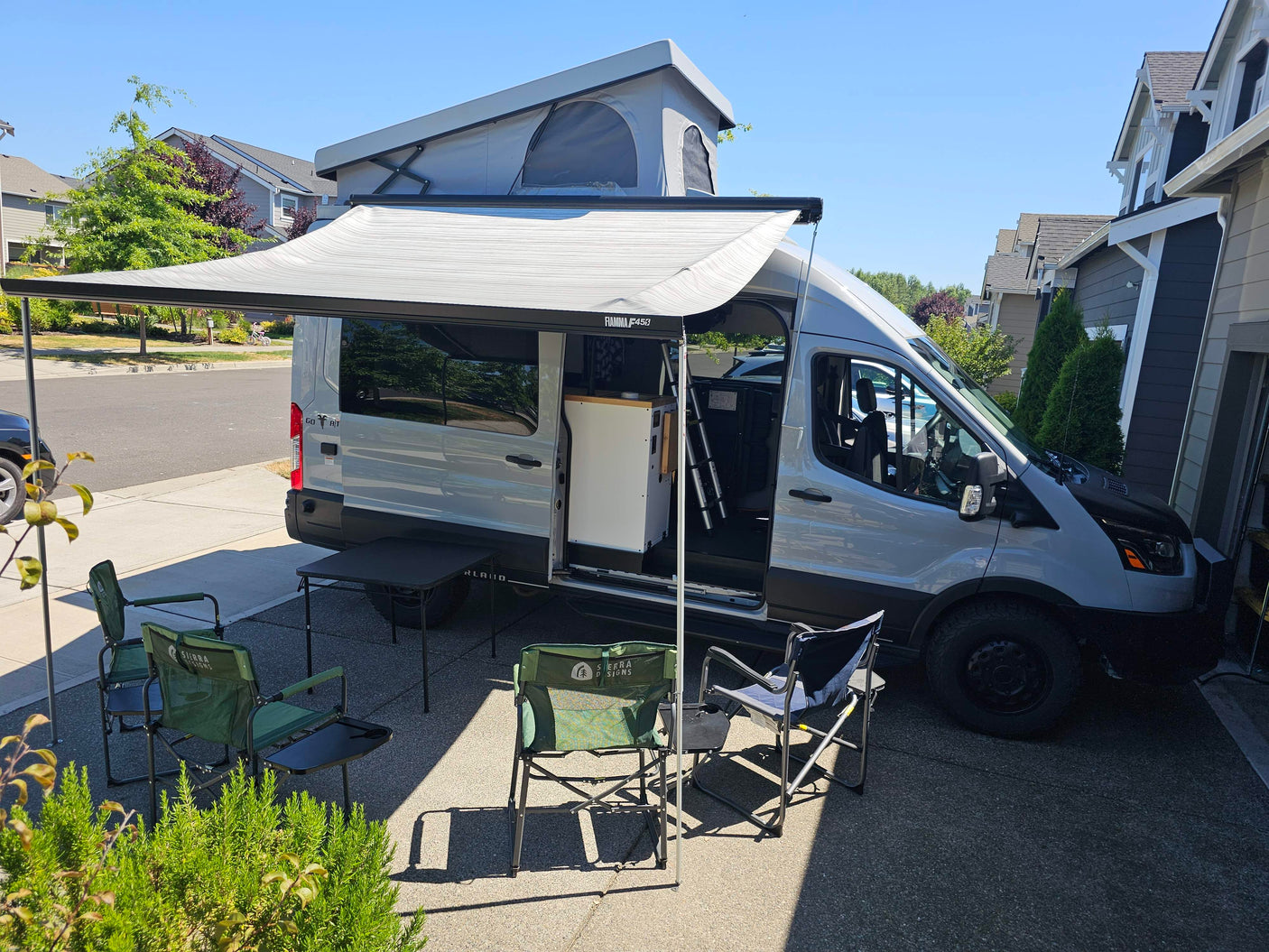 Gray RV rental campervan with pop-top roof and extended awning, set up with camping chairs and table for outdoor comfort.