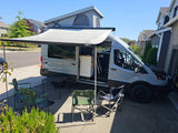 Gray RV rental campervan with pop-top roof and extended awning, set up with camping chairs and table for outdoor comfort.