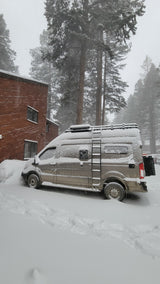 A snow-covered RV rental van is partially buried in deep snow, parked next to a building and tall trees.