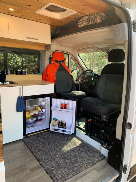 Inside a modern RV rental van: open fridge with food, compact kitchenette, driver's area, wood ceiling, and cozy rug.