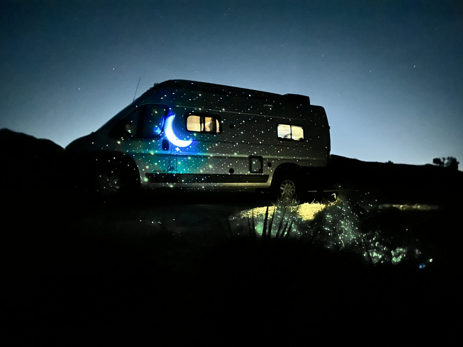 An RV glows with projected stars and a crescent moon under a dark night sky. Imagine this peaceful rv rental experience.