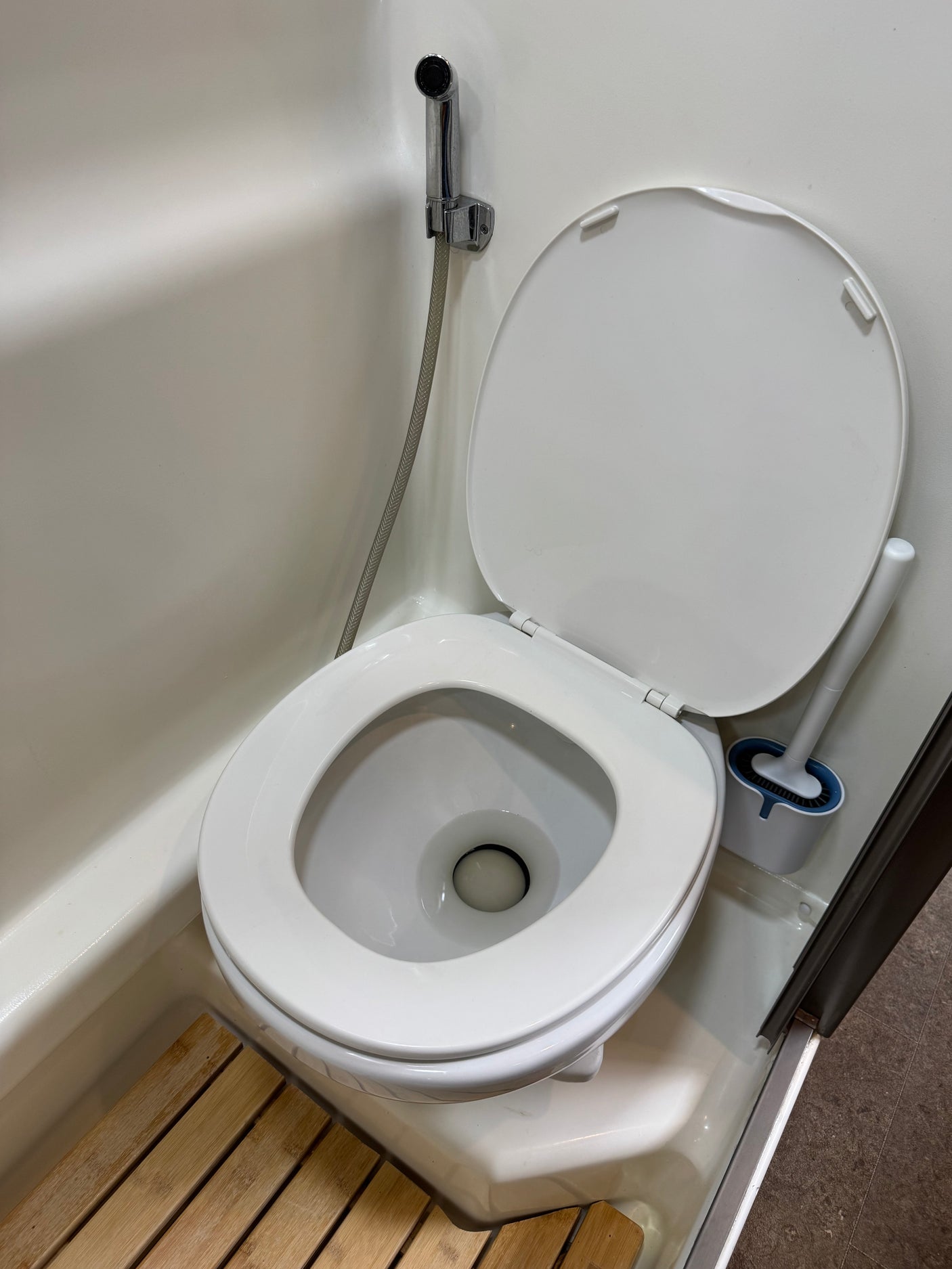 Compact RV rental bathroom featuring a white toilet with bidet spray, toilet brush, and a wooden slatted shower mat.