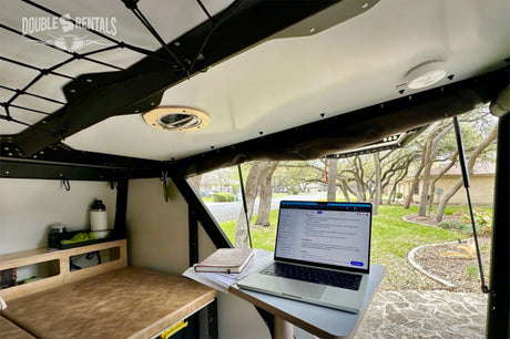 Double S RV rental offers a mobile office setup: laptop, bed, and a green outdoor view from the camper.
