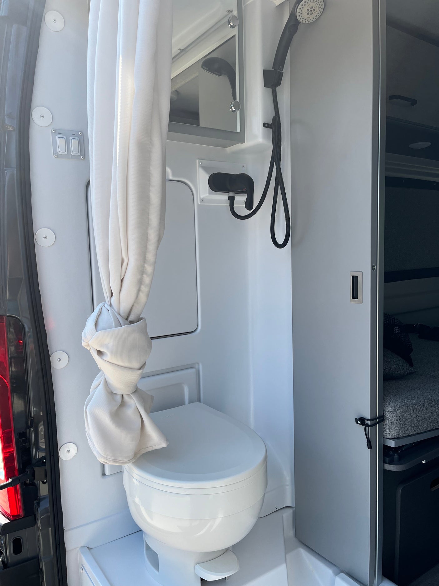 An RV rental's compact wet bath showing a toilet, shower, mirror, and light/fan switches, optimizing space for travel.