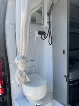 An RV rental's compact wet bath showing a toilet, shower, mirror, and light/fan switches, optimizing space for travel.
