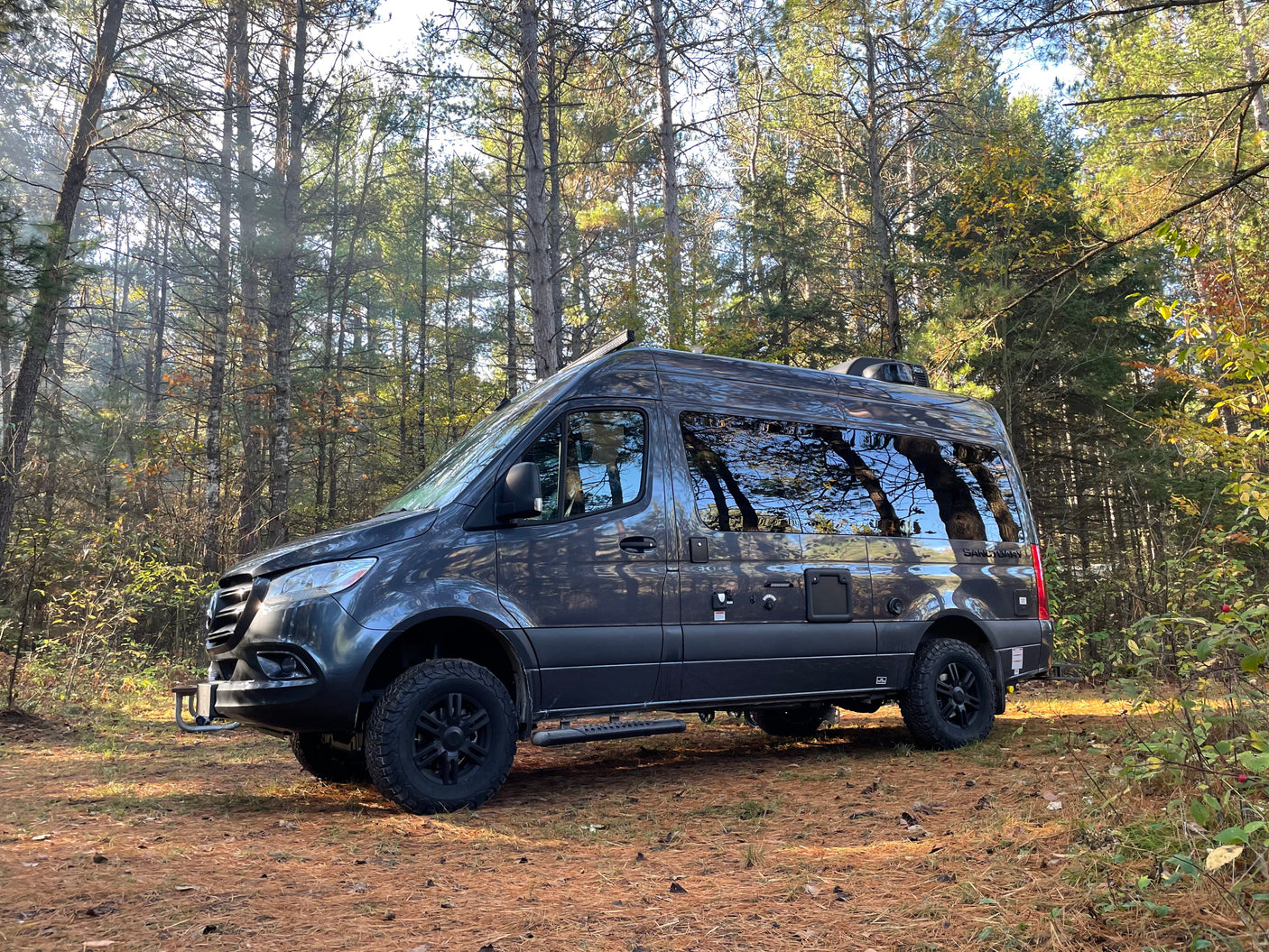 Dark grey camper van rv rental nestled among tall, sunlit pine trees. Ideal for a serene forest getaway.