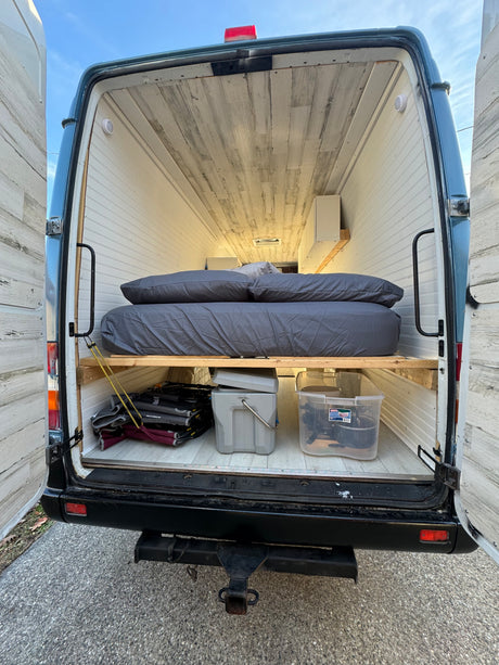 This RV rental's camper van interior features a raised bed, under-bed storage, and light wood-look paneling.