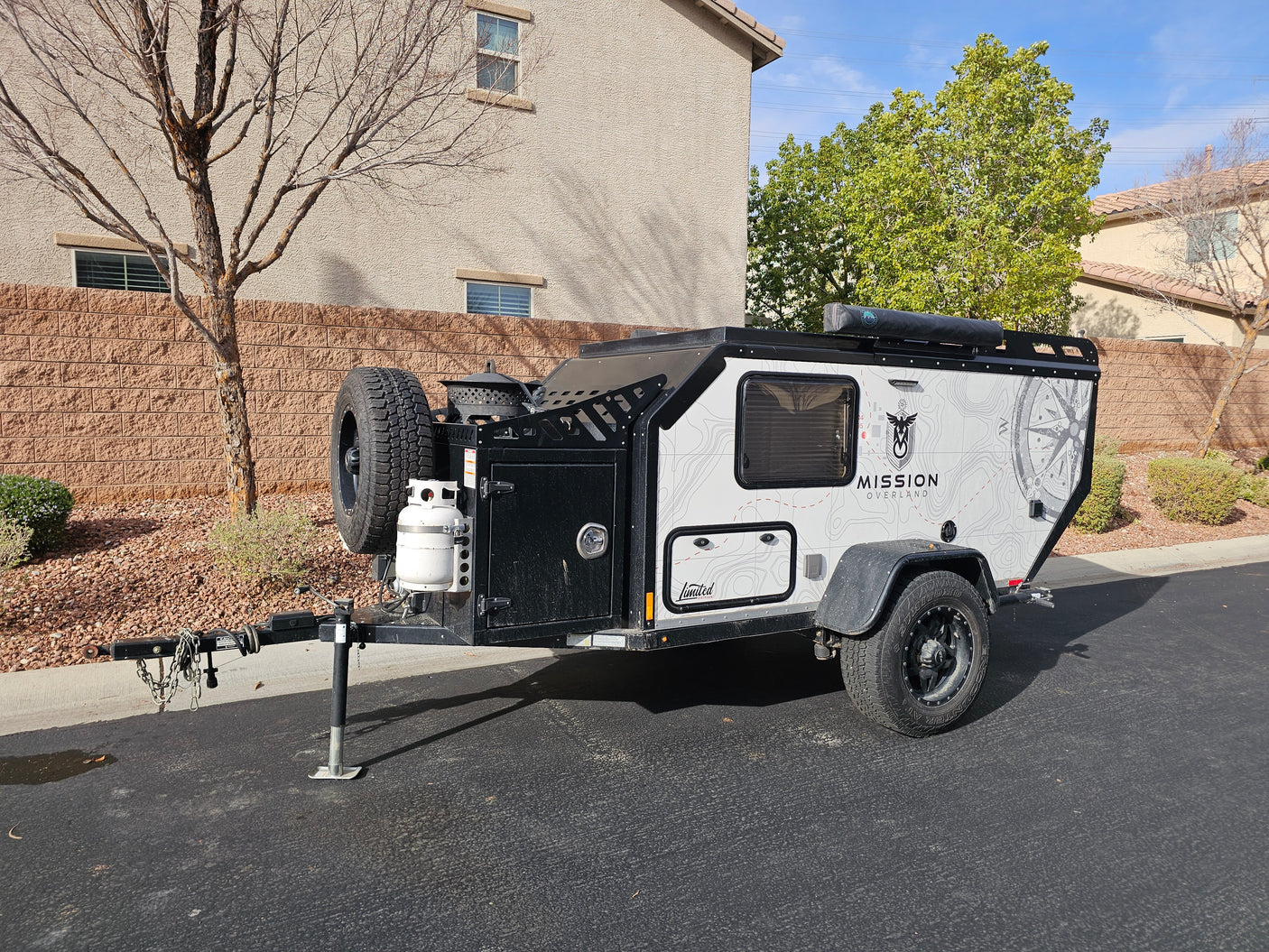 A Mission Overland off-road camping trailer, an excellent RV rental option, is parked outdoors with a spare tire & propane tank.
