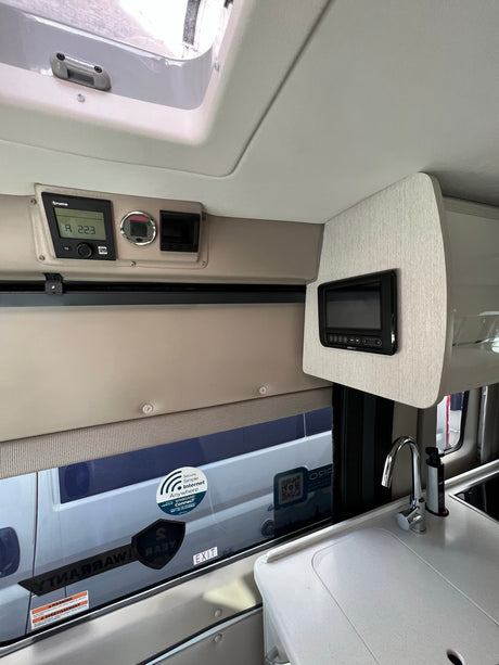 Inside an RV rental: Controls for heating and systems, a compact kitchen with sink, and window showing another vehicle, offering comfort for travel.
