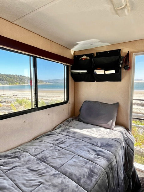 RV rental interior: A cozy gray bed, wall organizers, and a stunning beach view from the large window in this sleeping area.