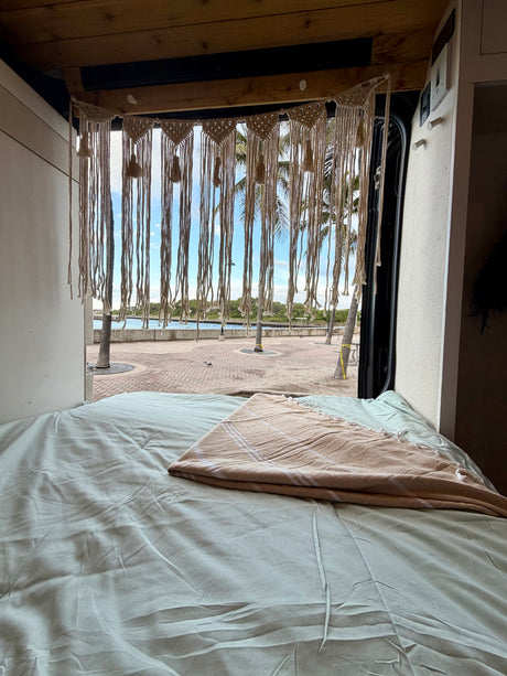 Cozy RV rental bed with a macrame curtain, offering a tropical waterfront view with palm trees. Ideal for van life.