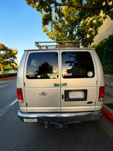 Silver Ford van, possibly an RV rental, with a roof rack and adventure stickers like "KIRKWOOD" and "MOUNTAIN BIKER" on its rear.
