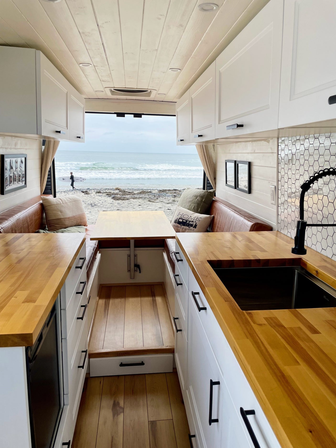 This rv rental shows a bright camper interior with a kitchen, dining area, and an ocean view at a beach with a surfer.