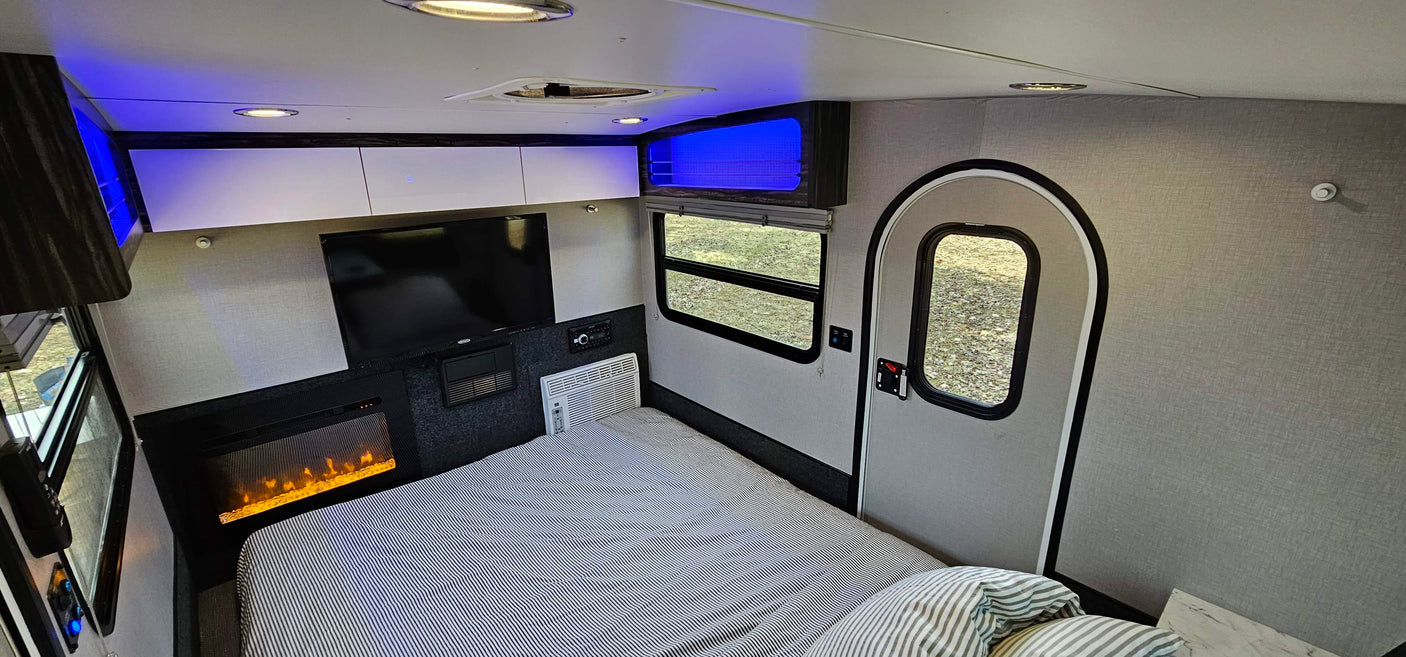 Step inside this cozy RV rental bedroom, complete with a striped bed, TV, electric fireplace, AC, and blue accent lighting.