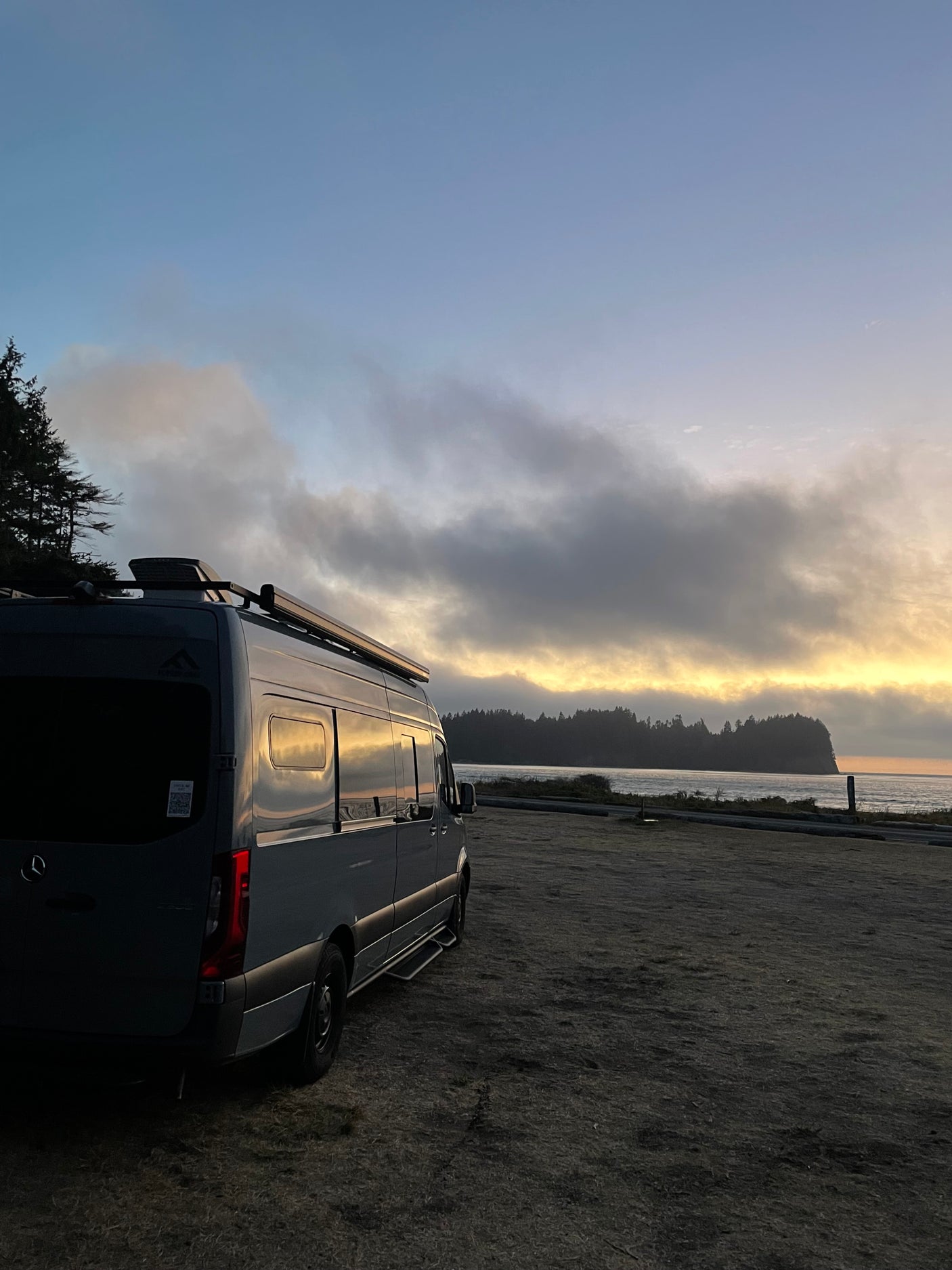 Dark grey Sprinter RV rental parked at sunset by the ocean, with a distant forested headland. Ideal for a coastal road trip.