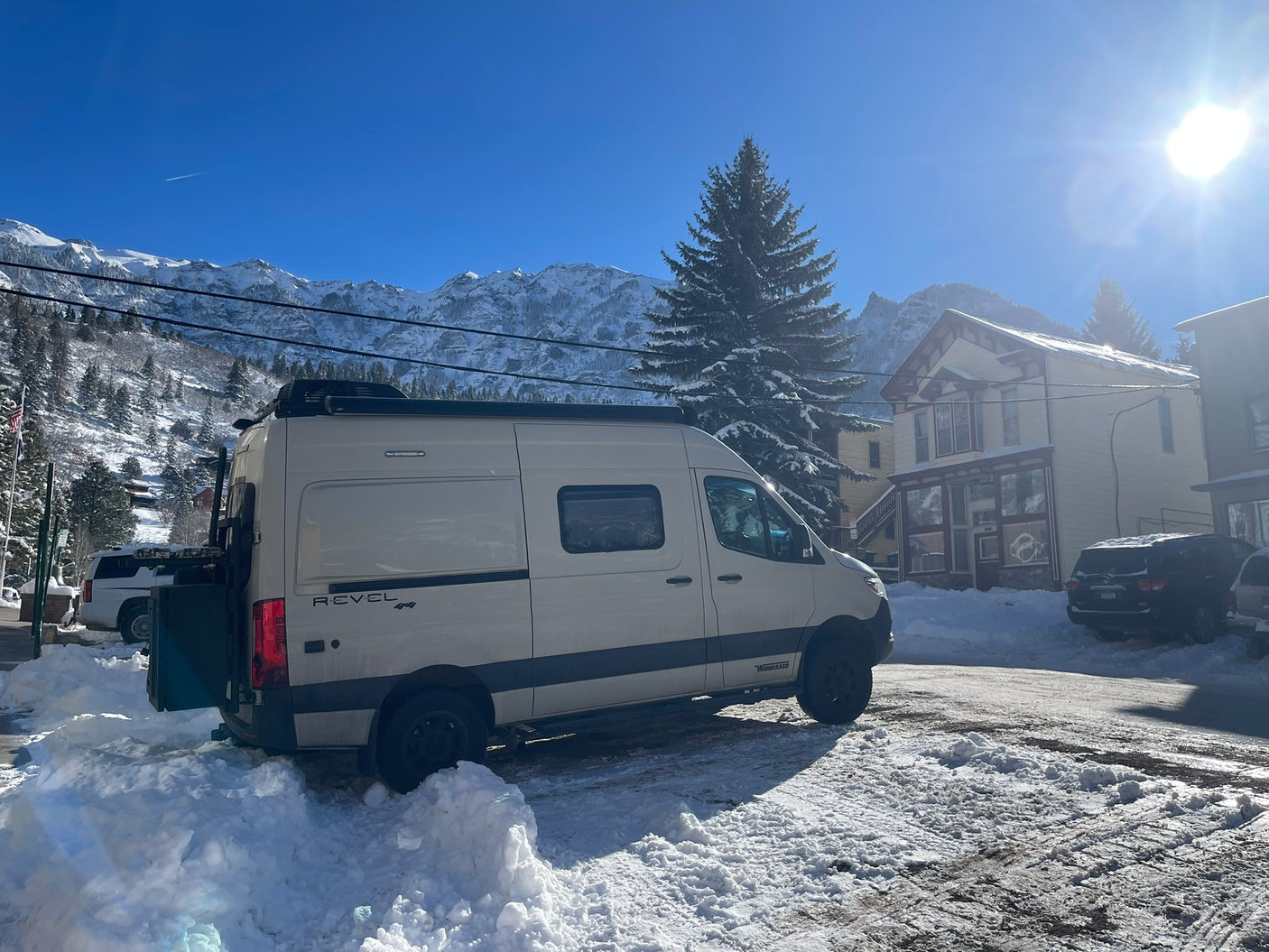 A light-colored Winnebago Revel rv rental van parked in fresh snow, with majestic mountains & bright sun overhead.