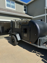 Dark, textured off-road teardrop camper, an RV rental option, parked on a sunny driveway by a house.