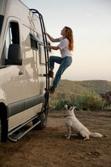 Woman ascends an RV rental camper van ladder, her dog watches below. Perfect for your next outdoor adventure or road trip.
