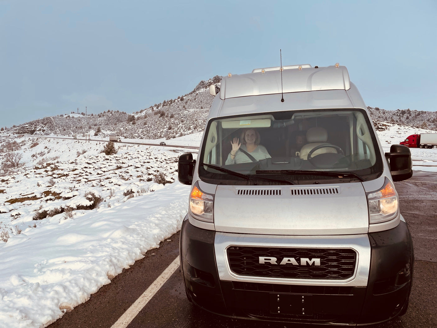 Winter adventure in an RV rental! A person waves from a Ram ProMaster van, parked amidst snow-covered hills.