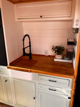 This modern RV rental kitchenette features light wood countertops, a white farmhouse sink, and a black faucet, with pale green cabinets below.