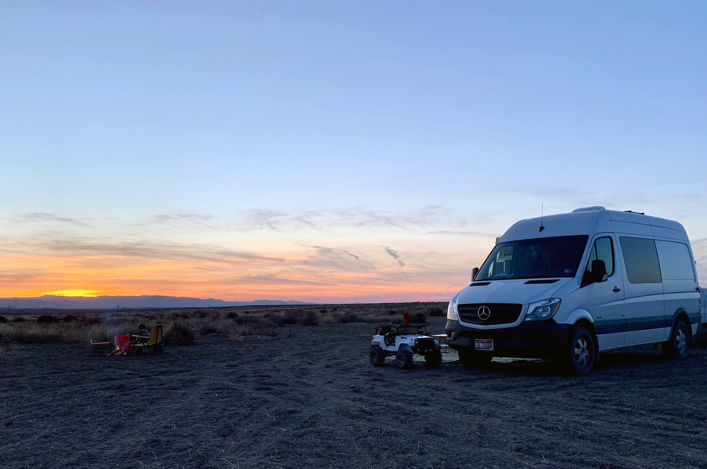 Enjoying an RV rental sunset! A camper van and toy Jeep by a campfire with a person, against a colorful sky in an open landscape.
