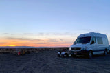 Enjoying an RV rental sunset! A camper van and toy Jeep by a campfire with a person, against a colorful sky in an open landscape.