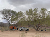 An SUV tows an orange teardrop RV rental, parked on sandy terrain under large trees, ideal for outdoor camping.