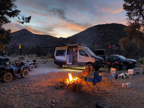 RV rental camp at dusk: a cozy campfire, camper van, truck, ATVs, and dogs in a mountain setting.