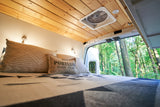 Relaxing RV rental bed with a 'Portland' pillow, wooden ceiling, and serene forest view from the open rear doors.