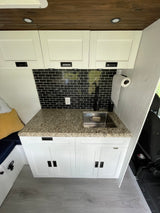 Modern RV rental kitchen: white cabinets, granite countertop, black subway tile backsplash, stainless sink, and wooden ceiling.