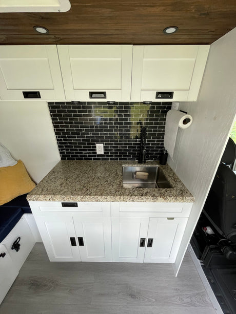 Modern RV rental kitchen: white cabinets, granite countertop, black subway tile backsplash, stainless sink, and wooden ceiling.