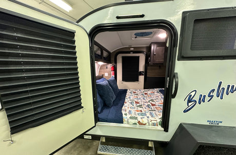 Step into this compact Bushwacker RV rental, showcasing its cozy interior bedroom with bed, storage, and open door.
