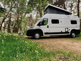 RV rental: A white pop-top camper van parked in a green, flowery forest clearing with a person relaxing.