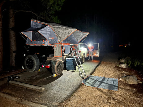 Night RV rental setup: an overlanding trailer with a rooftop tent beside an SUV at a campsite, illuminated by headlamp.