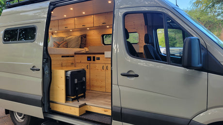 Light-colored RV rental camper van with open side door, showcasing a cozy, custom wood-paneled interior with bed and kitchen.