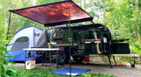 Scenic rv rental campsite: dark teardrop trailer, extended awning, blue tent, and outdoor table setup amidst green trees.