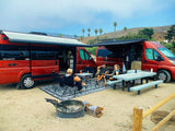 Two red RV rental vans at a sandy campsite. People relax outdoors with a picnic table, chairs, and a fire pit.