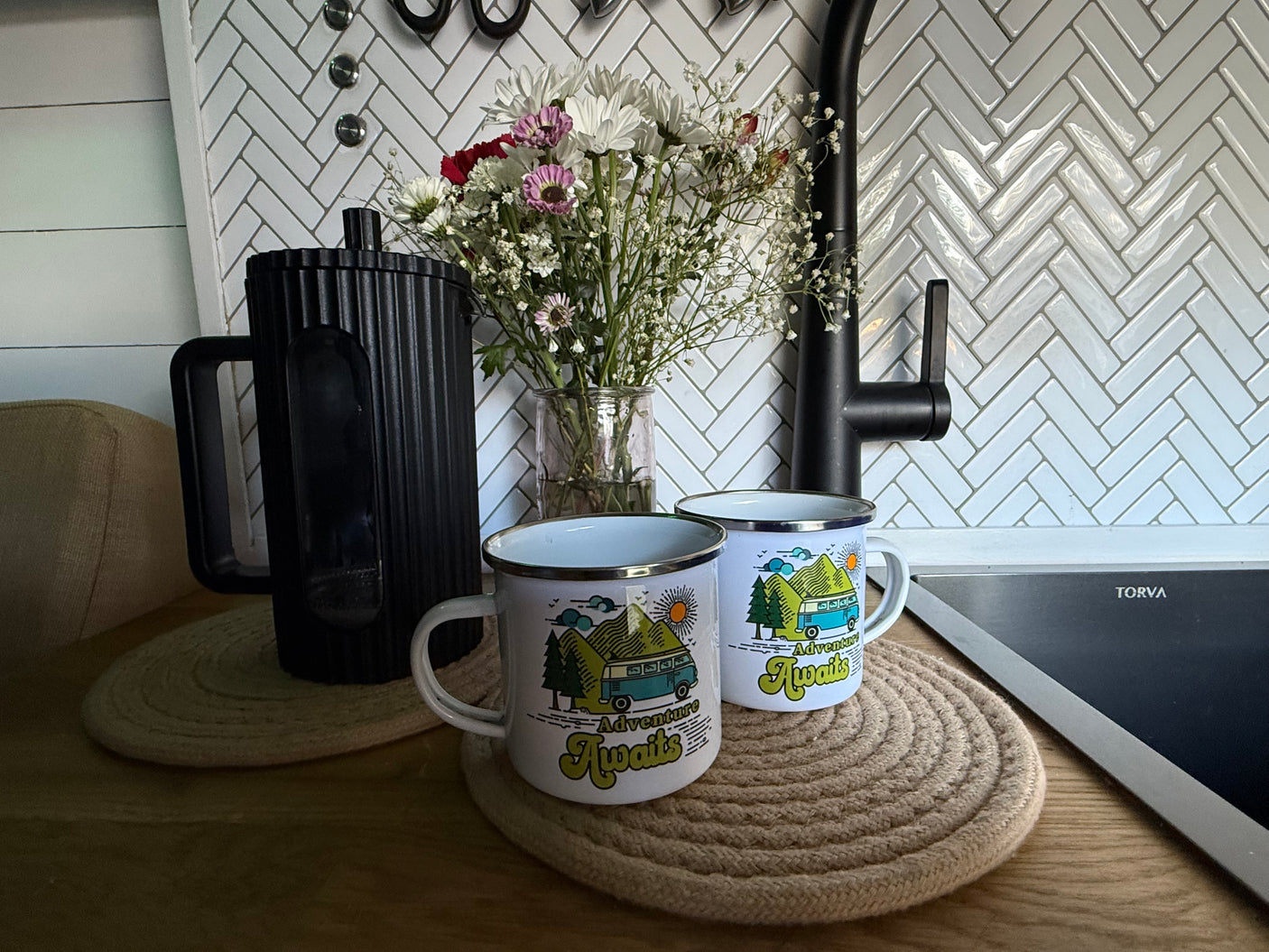 Cozy rv rental kitchen: 'Adventure Awaits' camper mugs, fresh flowers, & coffee pot on wood counter. Ready for travel!