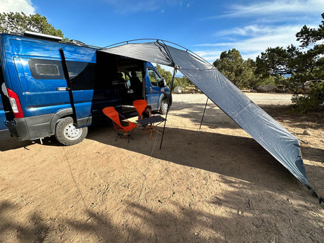 An RV rental camper van with an extended awning provides shade for chairs & a child on a sunny day.