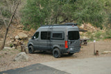 A rugged, dark gray Mercedes Sprinter camper van rv rental with off-road tires & roof rack, ready for adventure at a campsite.