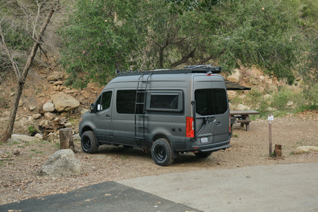 A rugged, dark gray Mercedes Sprinter camper van rv rental with off-road tires & roof rack, ready for adventure at a campsite.