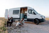 Couple enjoys drinks by their white Mercedes Sprinter rv rental camper van, interior visible at sunset.