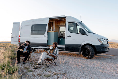 Couple enjoys drinks by their white Mercedes Sprinter rv rental camper van, interior visible at sunset.